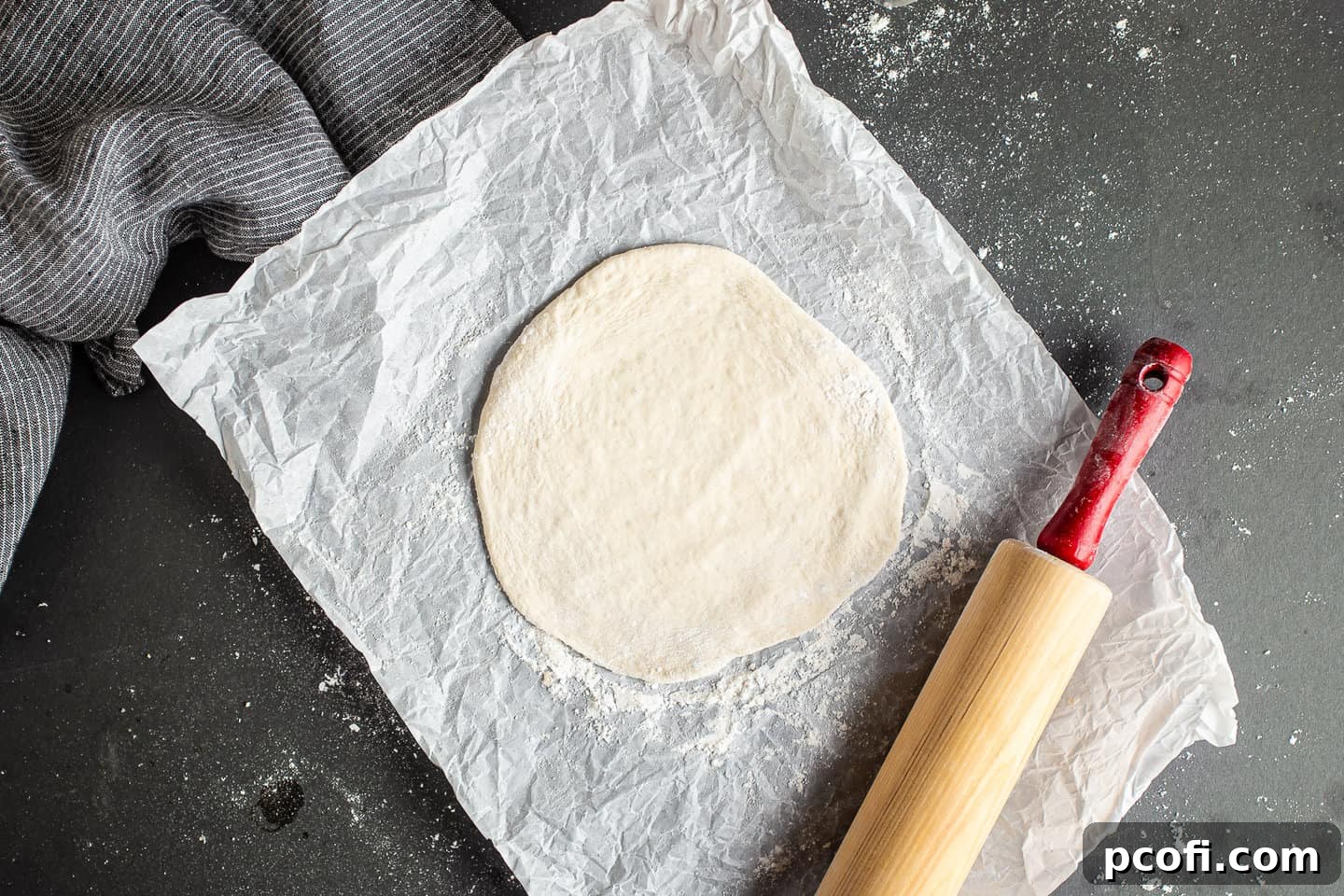 A person rolling out a portion of pita bread dough into a thin, round shape using a wooden rolling pin.