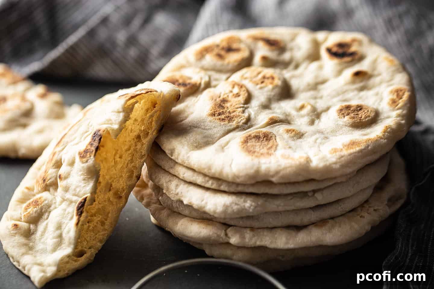A stack of warm, homemade pita bread, with one pita halved to showcase its soft interior, against a dark background.
