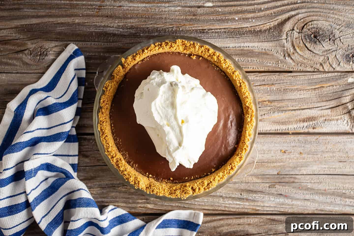 A freshly chilled chocolate pudding pie is being generously topped with homemade whipped cream, creating beautiful swirls and peaks.