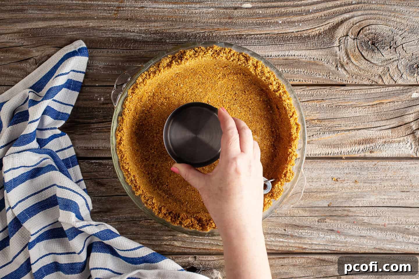 A measuring cup is used to firmly press the graham cracker mixture into the bottom and up the sides of a pie dish, forming an even crust.