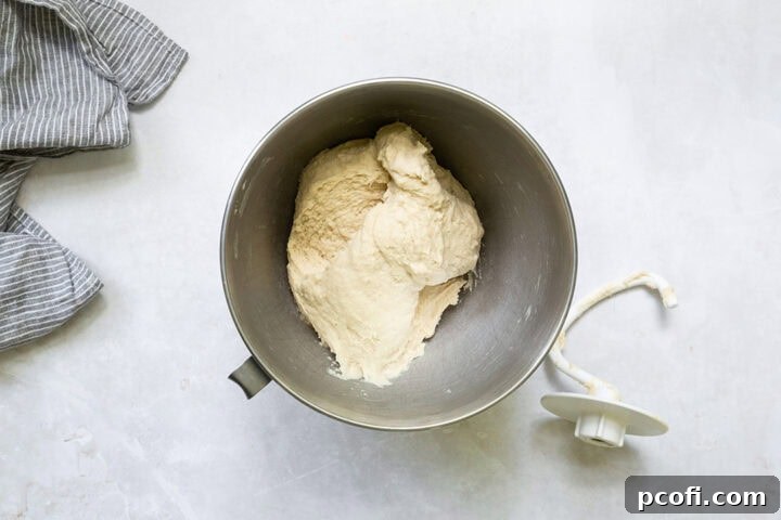 English muffin dough mixing in a stand mixer bowl with the dough hook attachment.