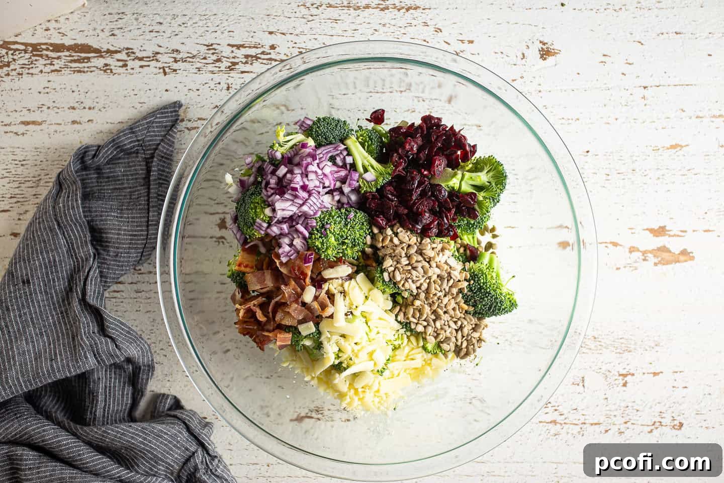 All the vibrant ingredients for broccoli salad, including florets, bacon, cheese, dried fruit, and seeds, are gathered in a large glass bowl, ready for tossing.