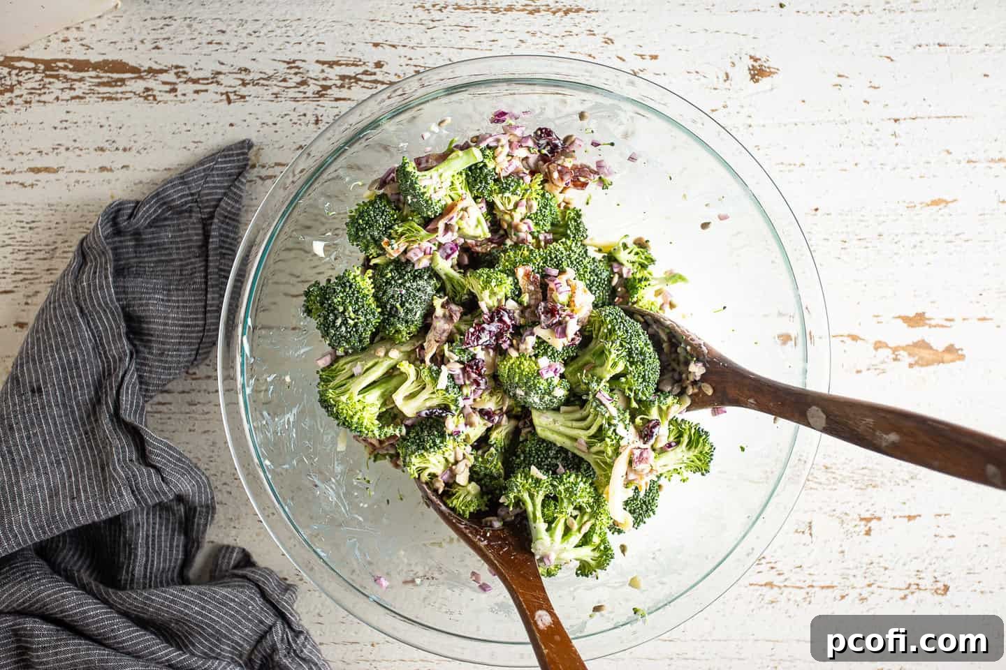 A pair of serving spoons gently tossing the colorful broccoli salad, ensuring every ingredient is evenly coated in the creamy dressing for a perfect mix.