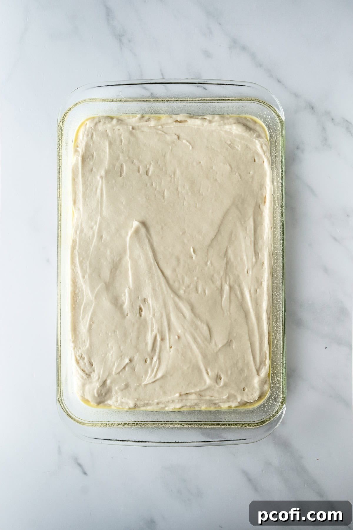 Vanilla cake batter poured into a greased 9x13 inch baking dish, ready for the oven, set on a white table.