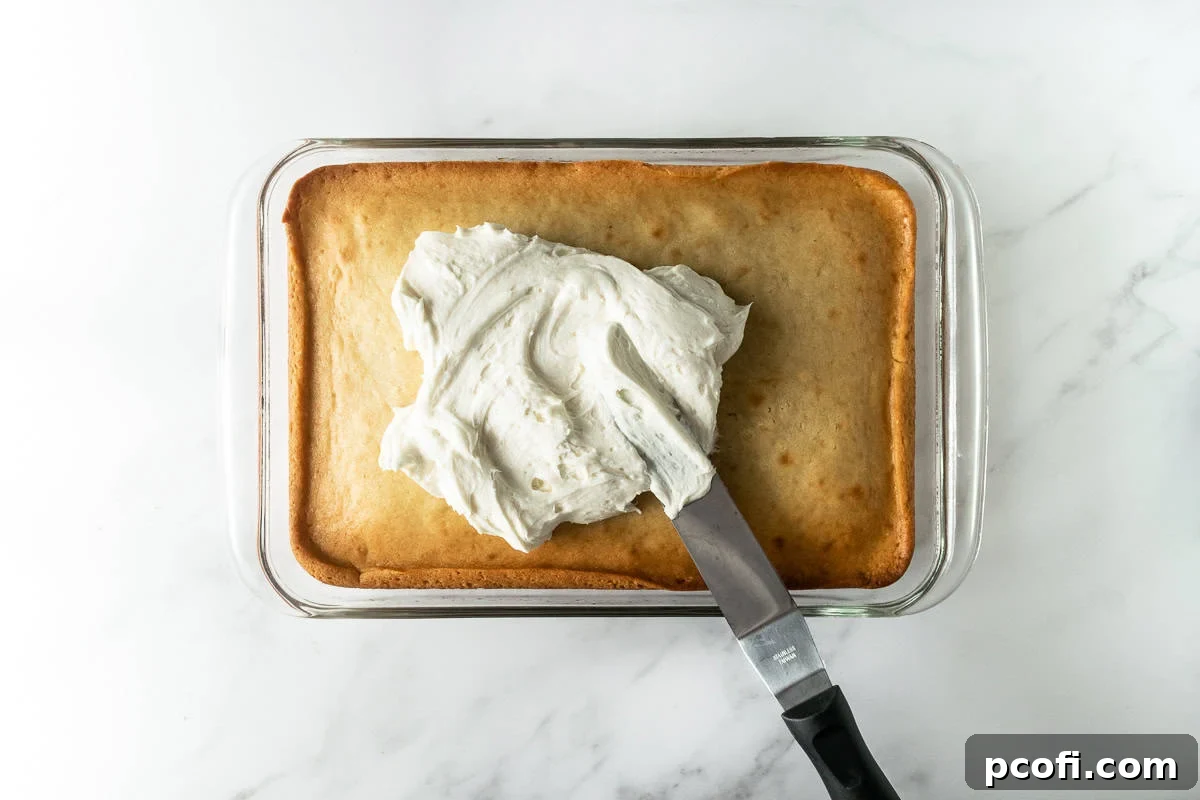 A vanilla cake in a baking pan, partially covered with creamy vanilla frosting, with an offset spatula resting beside it on a white surface.