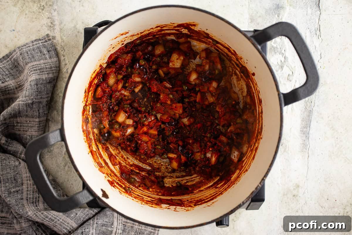 The vibrant tomato paste, olives, and capers cooking together in the pot, beginning to caramelize and develop a rich, brown 'fond' on the bottom, intensifying the sauce's flavor.