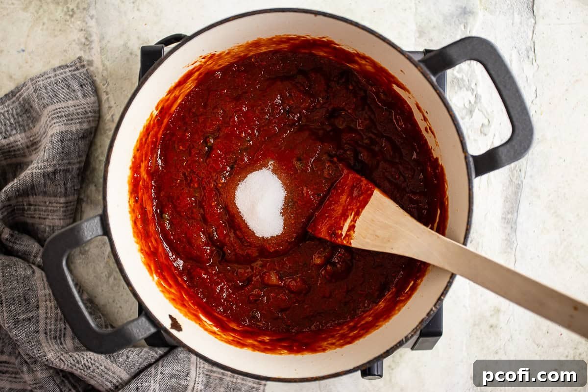 A hand adding a pinch of granulated sugar to a pot of simmering puttanesca sauce, illustrating the step to balance flavors.