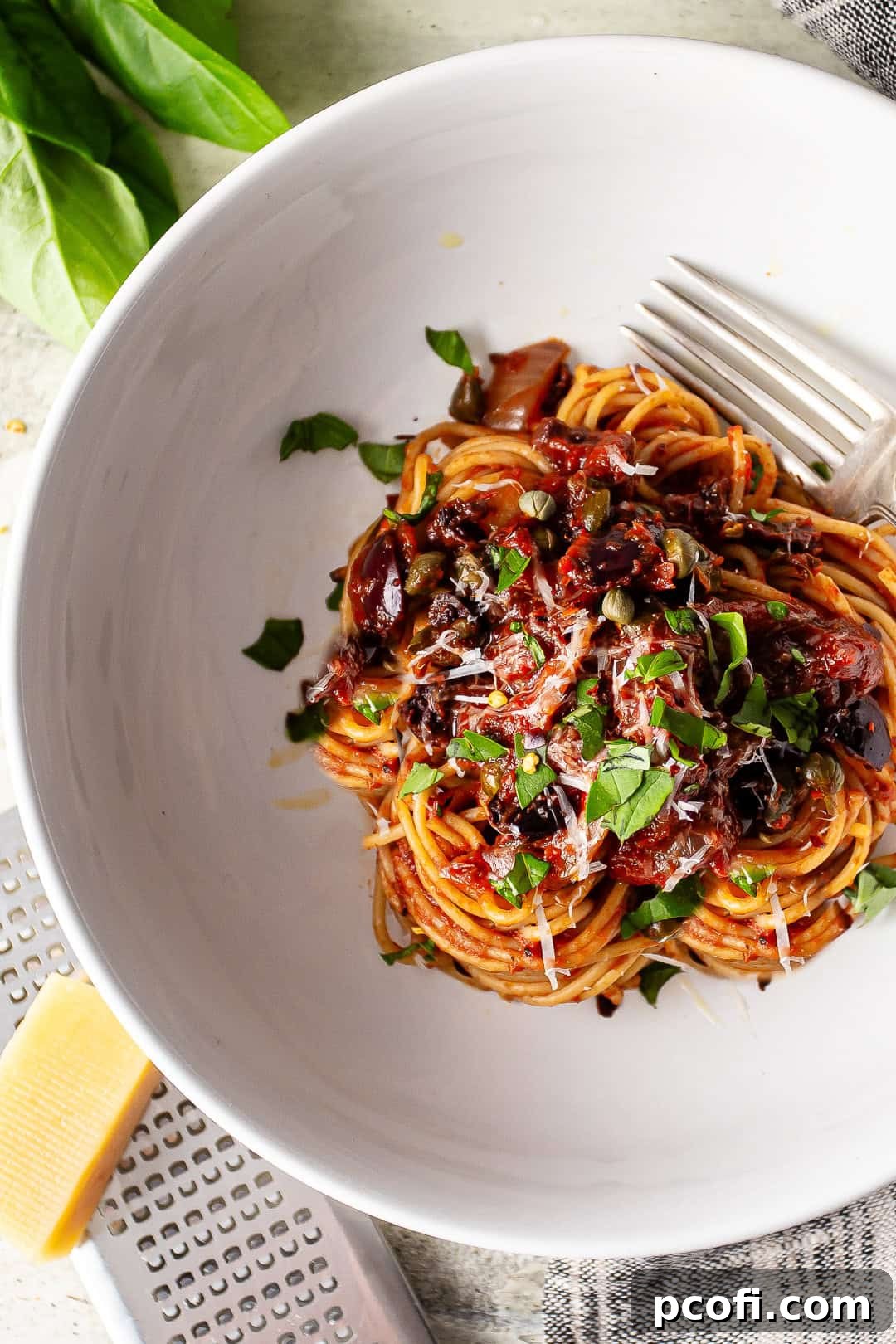 Puttanesca sauce served over thin spaghetti with fresh basil leaves and grated parmesan cheese in the background, showcasing the vibrant colors and textures of the dish.