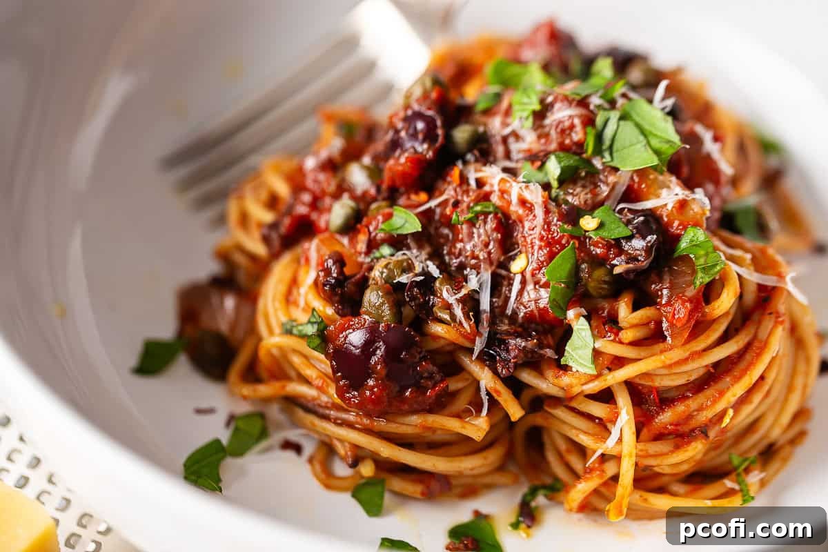 Close-up image of a vibrant spaghetti puttanesca recipe, highlighting the chunky black olives and small capers nestled within the rich red sauce, garnished with fresh herbs.