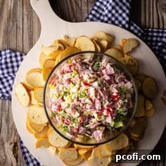 A bowl of Italian Hoagie Dip artfully arranged on a white wooden serving board, accompanied by an array of crunchy bagel chips.