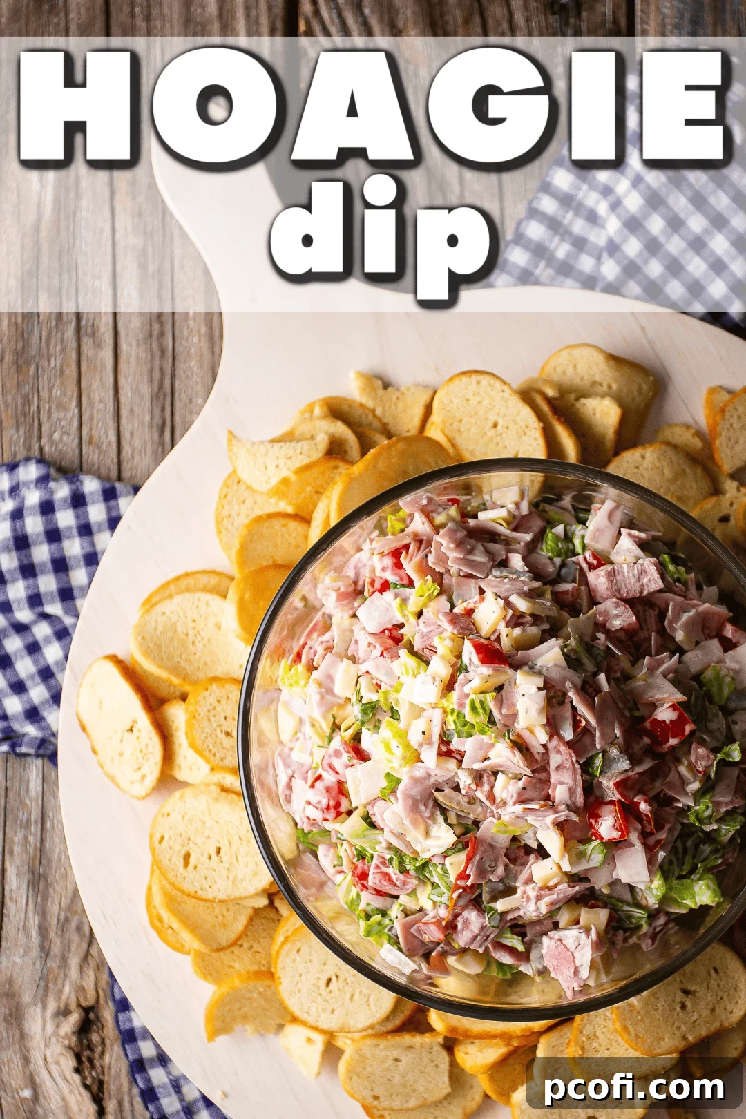 A glass bowl filled with Hoagie Dip, garnished and served on a wooden board alongside a generous pile of crispy bagel chips, ready for dipping.