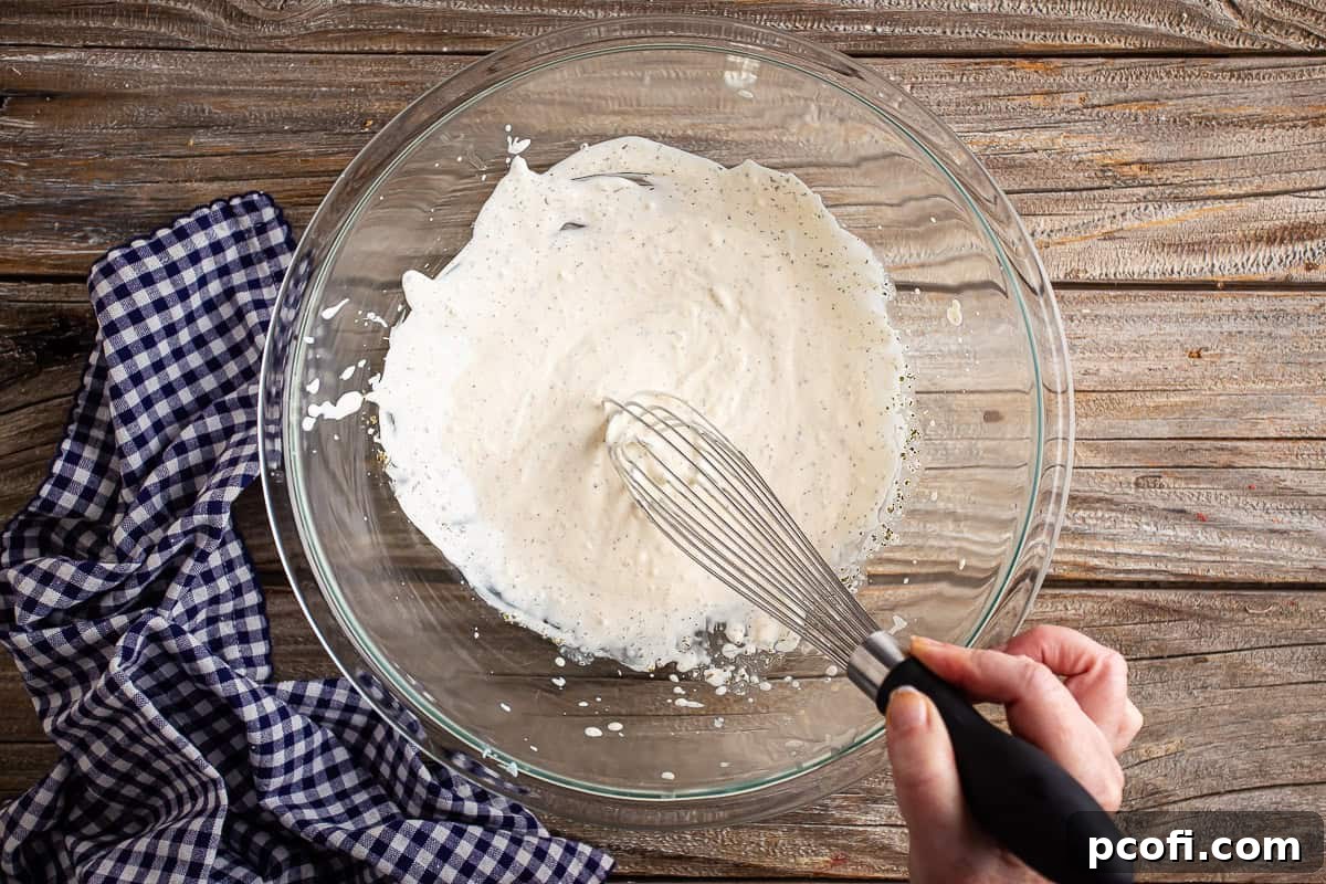 A whisk being used to combine mayonnaise, red wine vinegar, and Italian seasoning in a glass bowl, creating a smooth dressing.