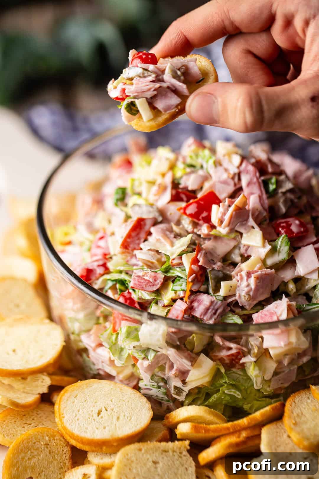 A hand scooping a generous portion of Italian Hoagie Dip onto a bagel chip, highlighting the dip's rich texture and fresh ingredients.