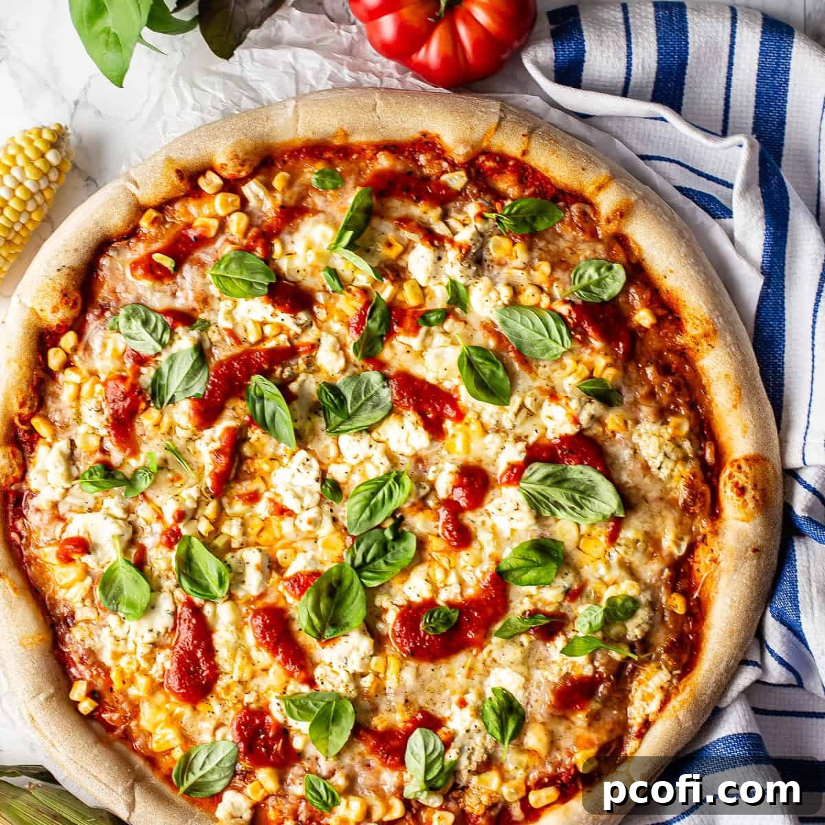 Overhead shot of a freshly baked corn pizza, adorned with vibrant green basil leaves, bright red tomato sauce, and dollops of creamy Boursin cheese, ready to be sliced.