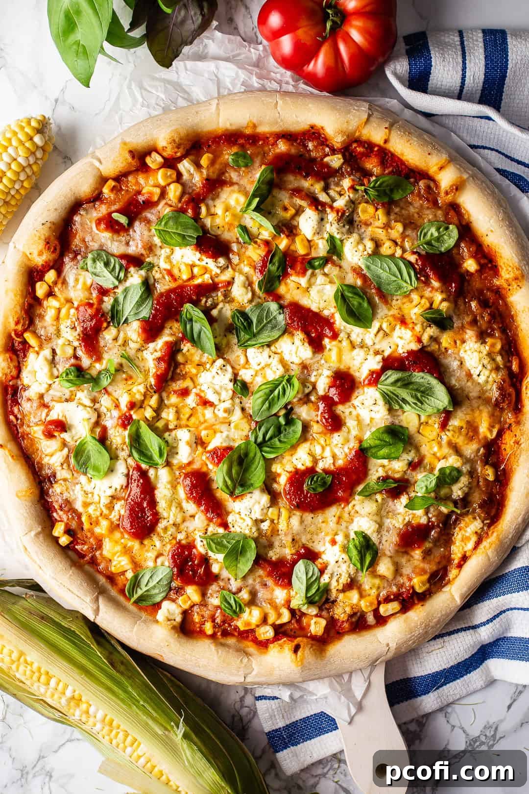 A vibrant homemade corn pizza, garnished with fresh basil leaves, sliced tomatoes, and creamy Boursin cheese, elegantly presented on a rustic board surrounded by fresh garden produce.