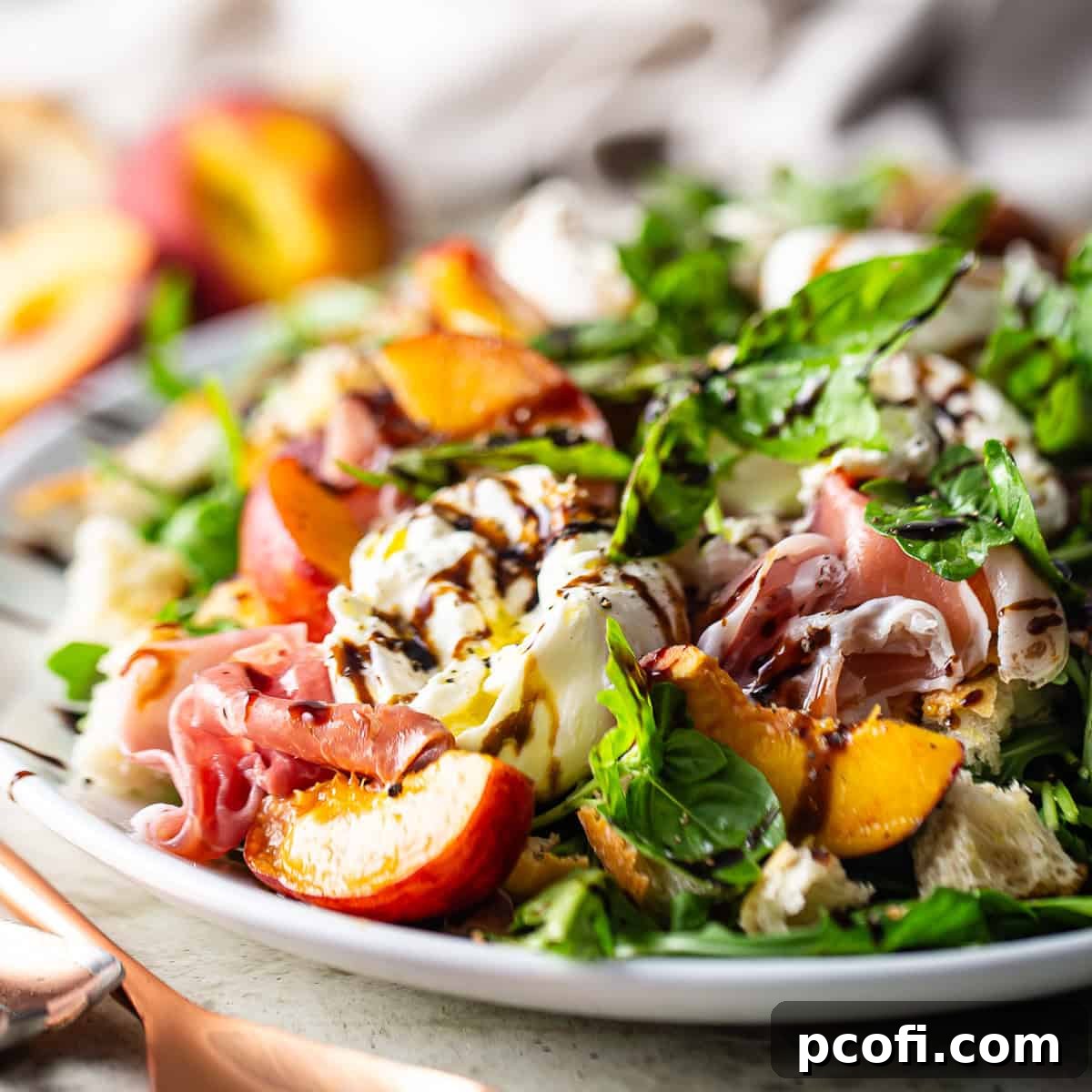 Peach salad presented on a white oval ceramic platter, showcasing fresh peaches, burrata, arugula, and prosciutto.