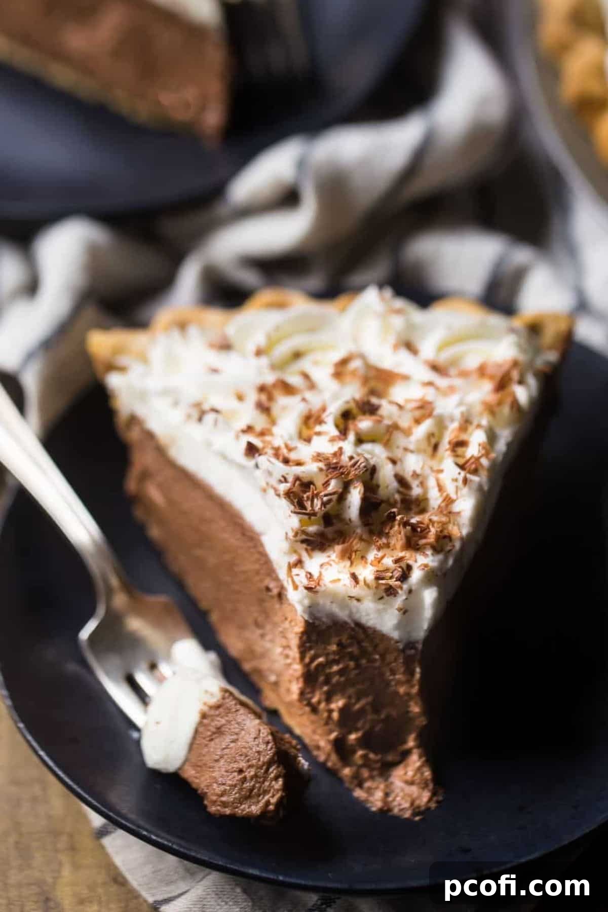 A perfectly sliced piece of chocolate cream pie, showcasing its rich layers of chocolate filling and whipped cream, presented on a white plate with a fork beside it.
