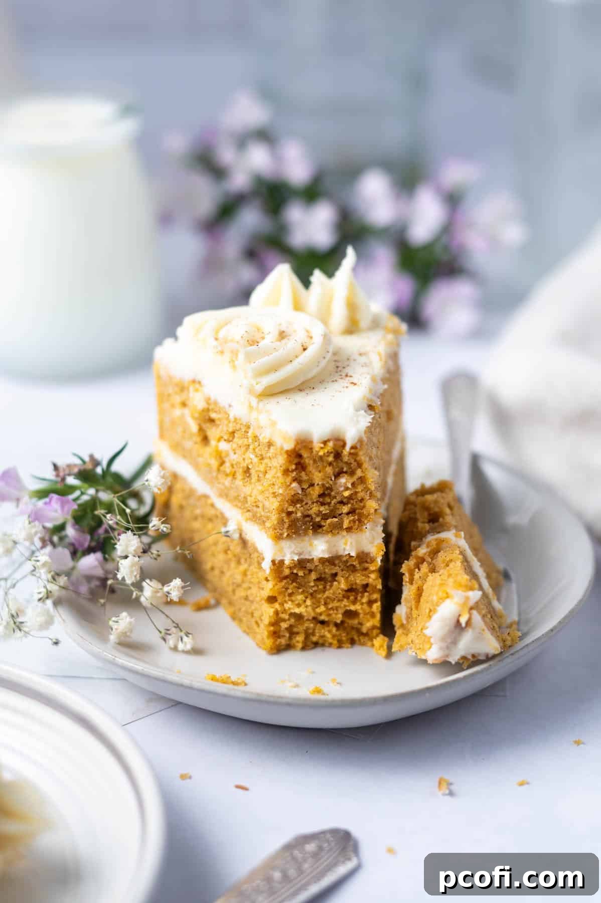 A beautiful slice of pumpkin layer cake with cream cheese frosting, elegantly presented on a plate next to a gold fork, with a glass of milk and autumnal flowers in the background.
