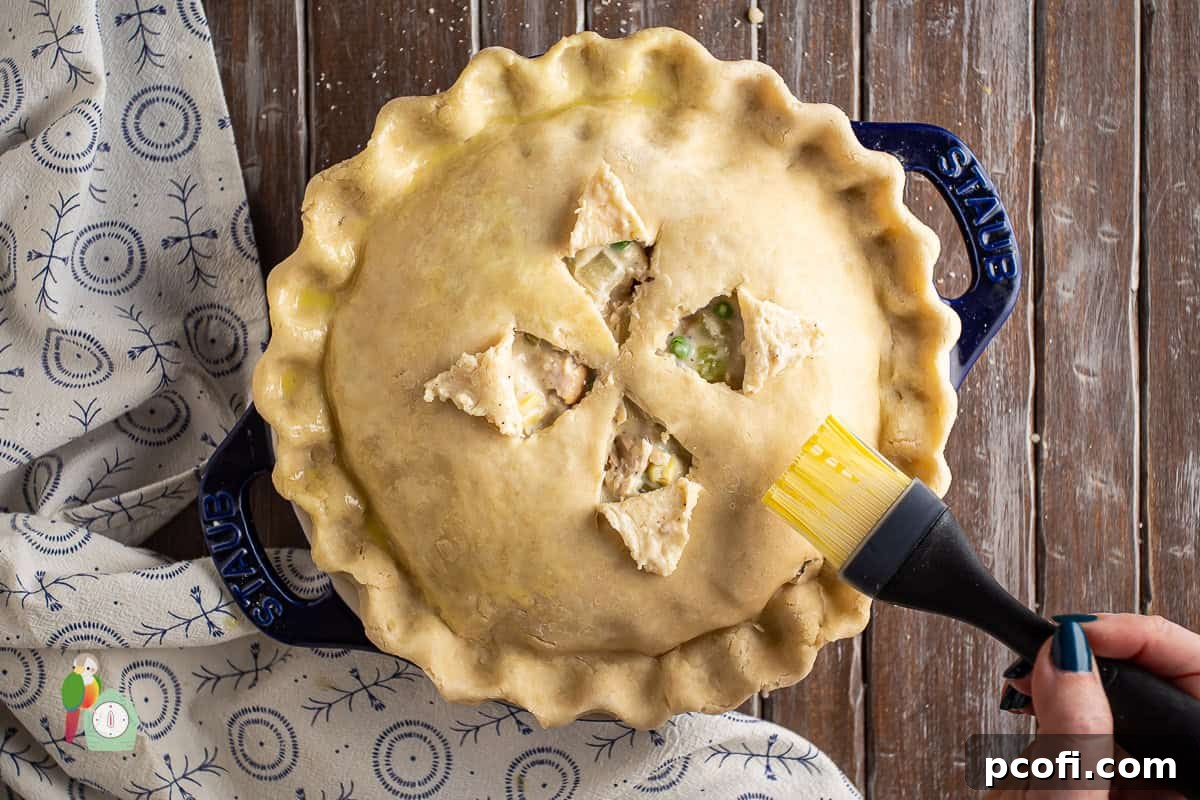 A hand brushing the top crust of an assembled turkey pot pie with an egg wash, ready for baking.