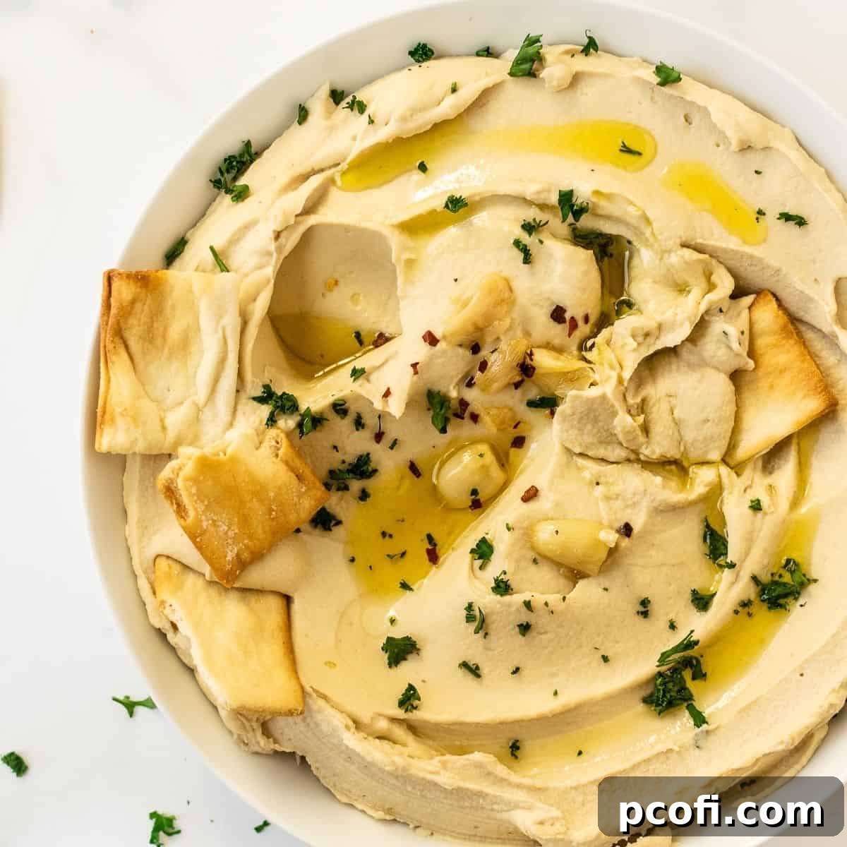 A smooth, flavorful hummus recipe presented in a white bowl, elegantly topped with fresh parsley, a drizzle of olive oil, and accompanied by pita chips.