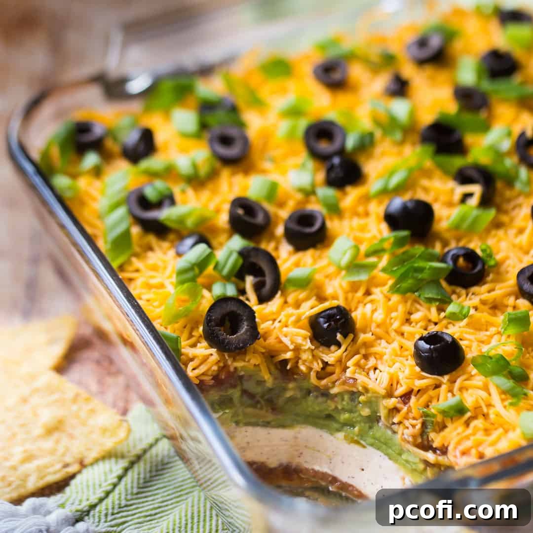 A square glass dish brimming with a vibrant 7-Layer Dip, perfectly garnished and ready to be scooped, alongside a green napkin.