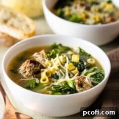 A beautifully presented bowl of Italian wedding soup, with visible mini meatballs, pasta, and greens, accompanied by crusty bread in the background.