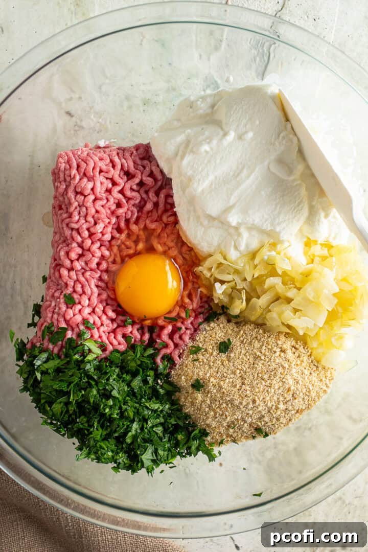 A large glass mixing bowl filled with ground meat, ricotta cheese, chopped onion, breadcrumbs, herbs, egg, and seasonings, ready to be mixed for meatballs.