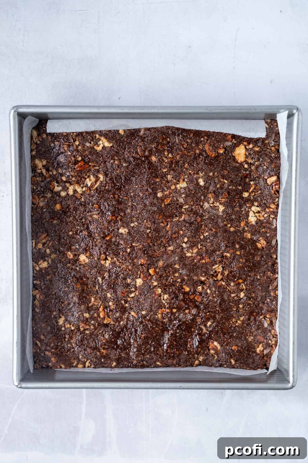 The chocolate, pecan, and coconut base mixture is pressed evenly into a parchment-lined baking pan, ready for chilling.