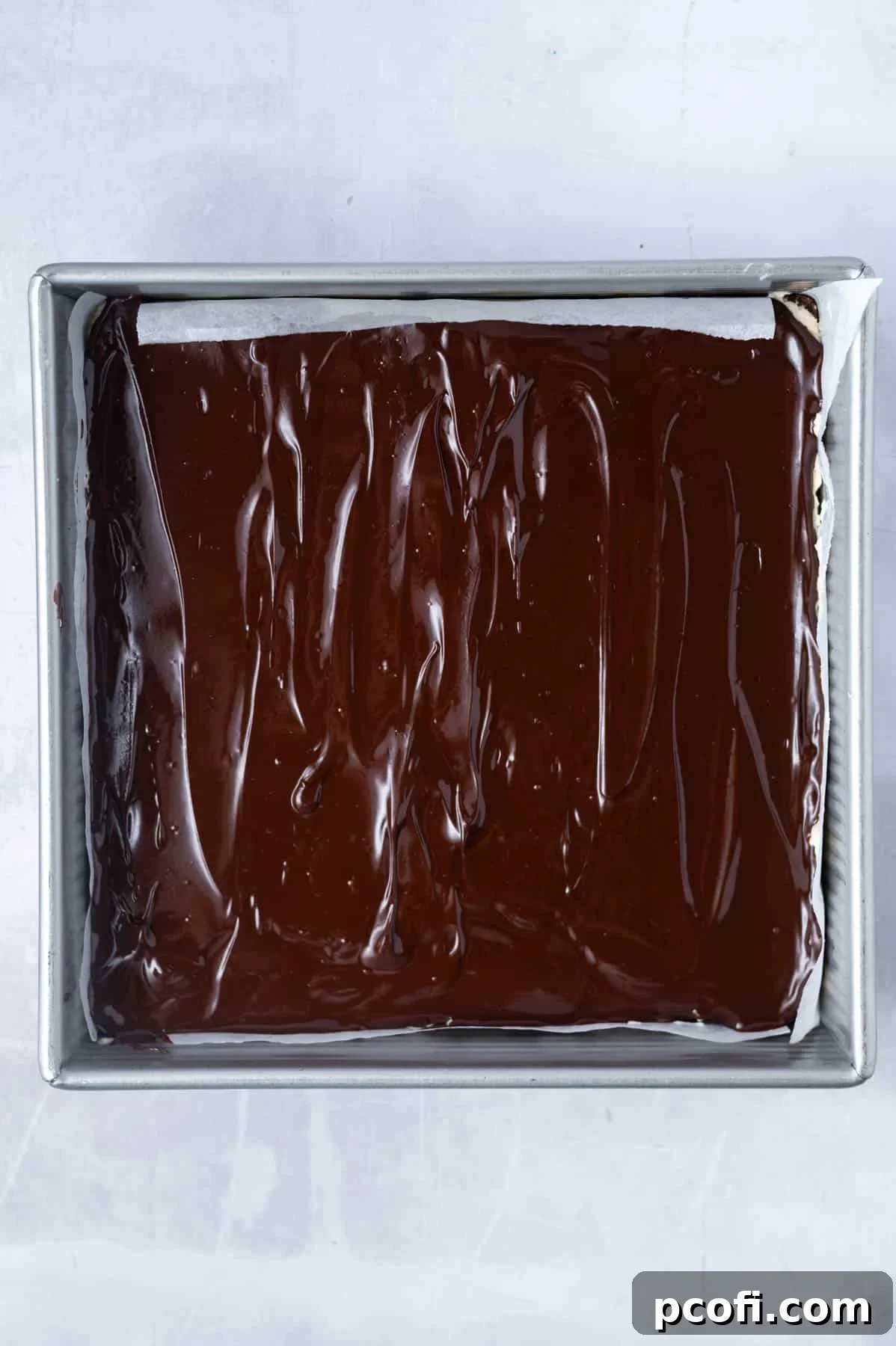 The smooth chocolate ganache layer is spread evenly over the chilled custard filling of the Nanaimo bars in a parchment-lined pan.