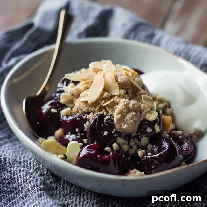 A vibrant bowl of no-bake cherry crumble, featuring glistening cooked cherries, a generous sprinkle of streusel and almonds, and a swirl of Greek yogurt on top.