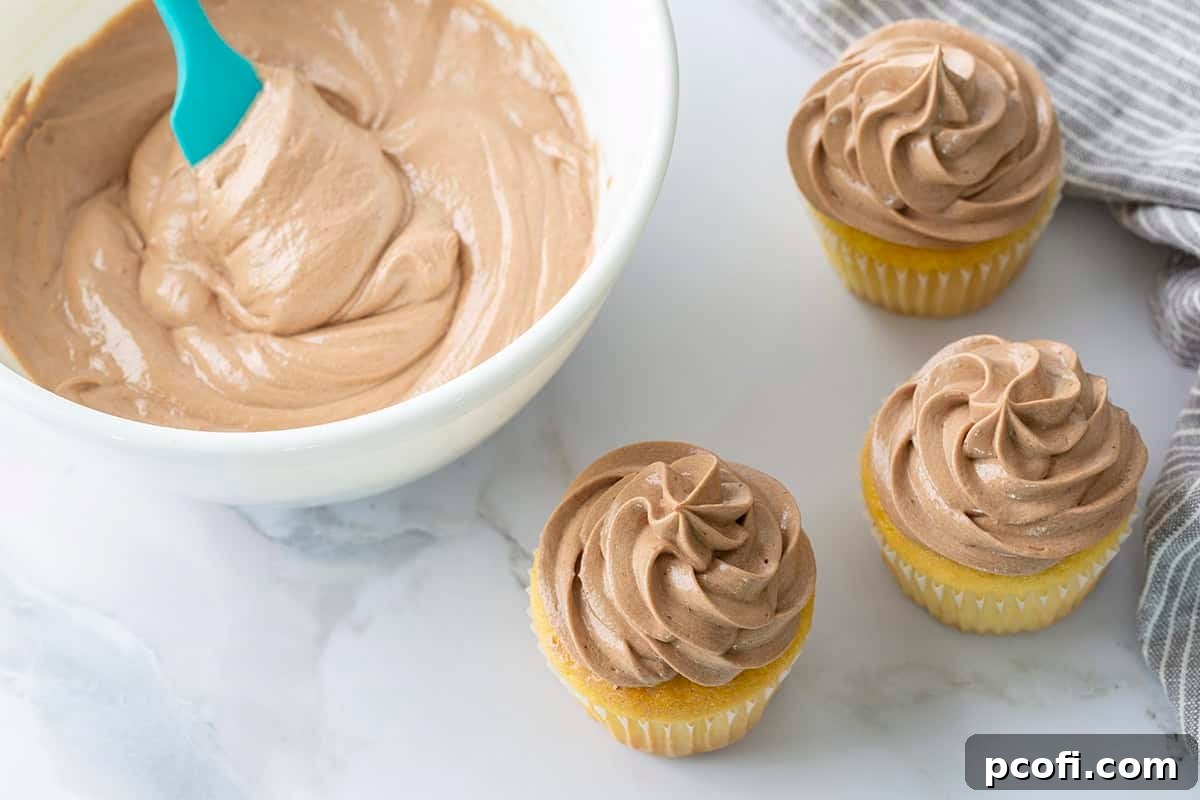 A generous bowl of smooth Chocolate Ermine Frosting next to beautifully frosted cupcakes, showcasing its perfect texture.