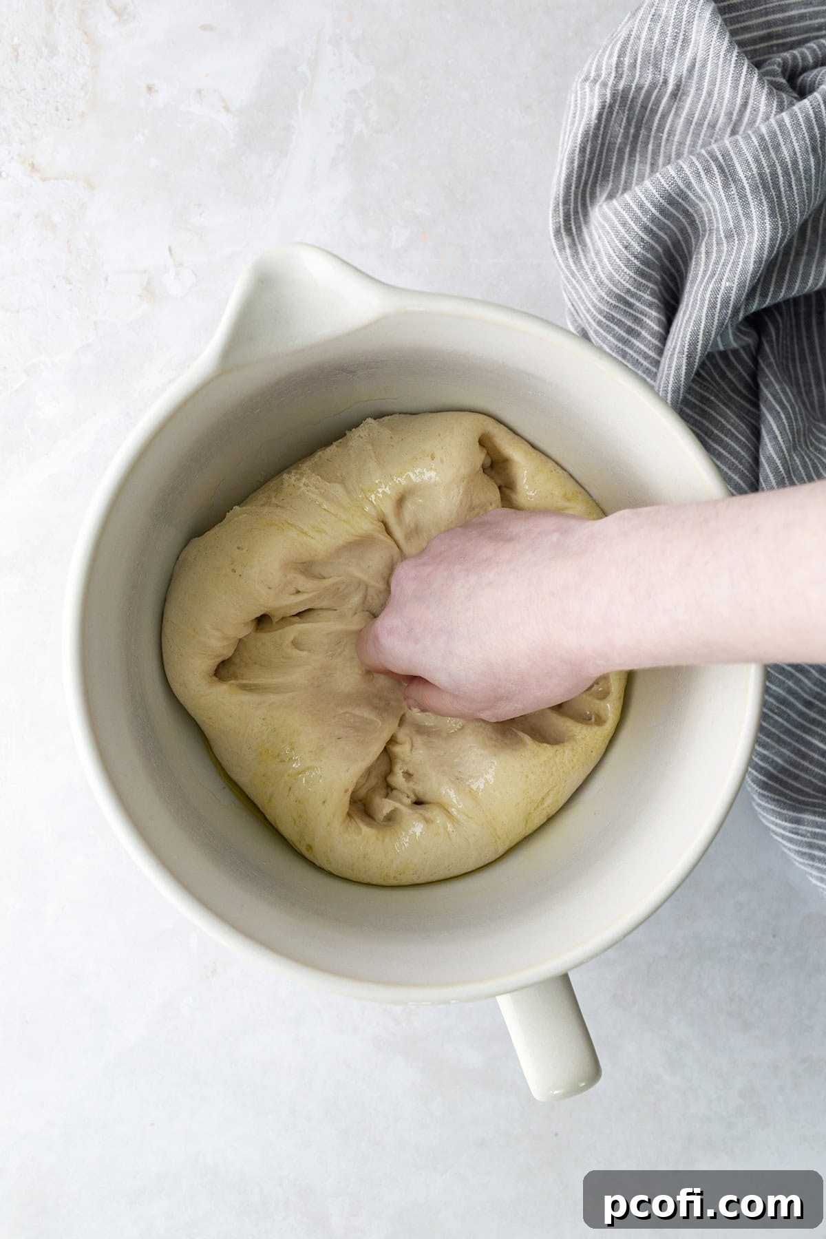 A hand gently punching down the risen potato bread dough in a bowl, releasing air and preparing it for shaping.