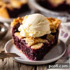 A slice of blackberry pie with ice cream on top and a fork near the dish.