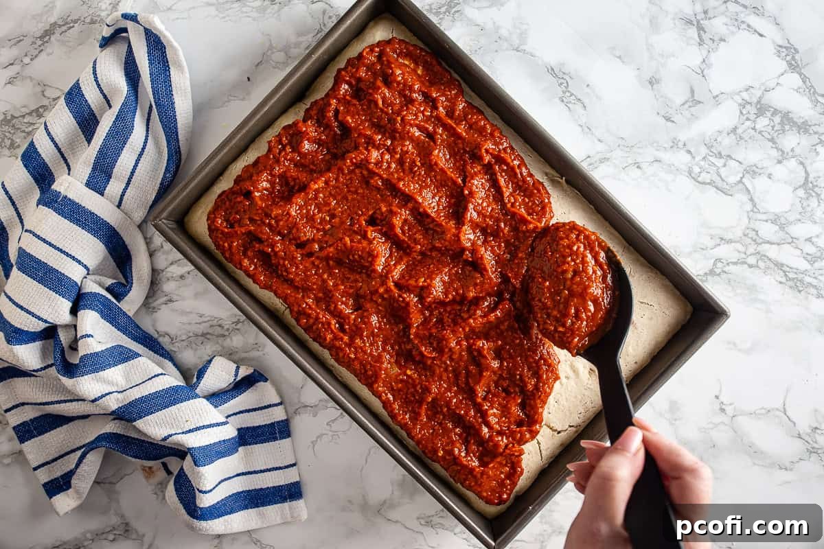 Tomato sauce being added to the top of a par-baked tomato pie pizza crust before being baked, showing a thick layer of red sauce.