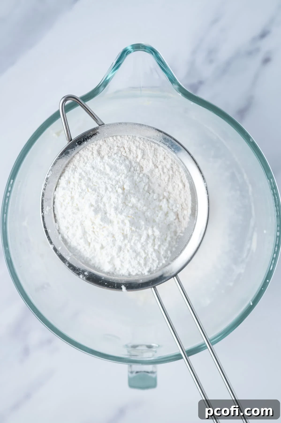 A sifter positioned over a mixing bowl, containing a blend of flour, cornstarch, and powdered sugar, ready to be incorporated into the creamy butter mixture.
