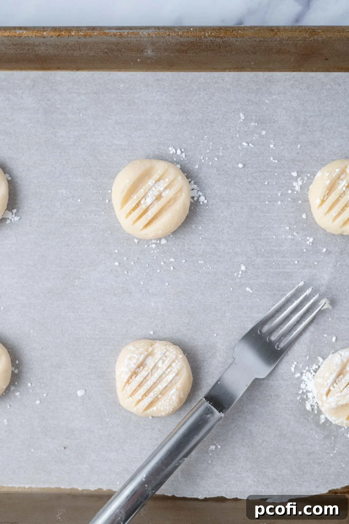 Freshly shaped cookie dough balls, lightly dusted with powdered sugar, awaiting baking on a parchment-lined pan, complete with delicate fork marks.