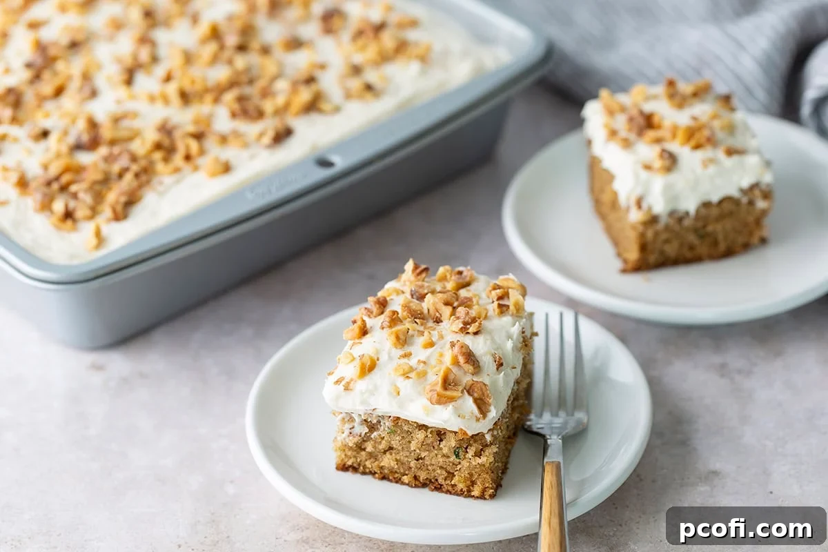 A perfectly sliced piece of zucchini cake, topped with cream cheese frosting and walnuts, resting on a white plate with a fork.