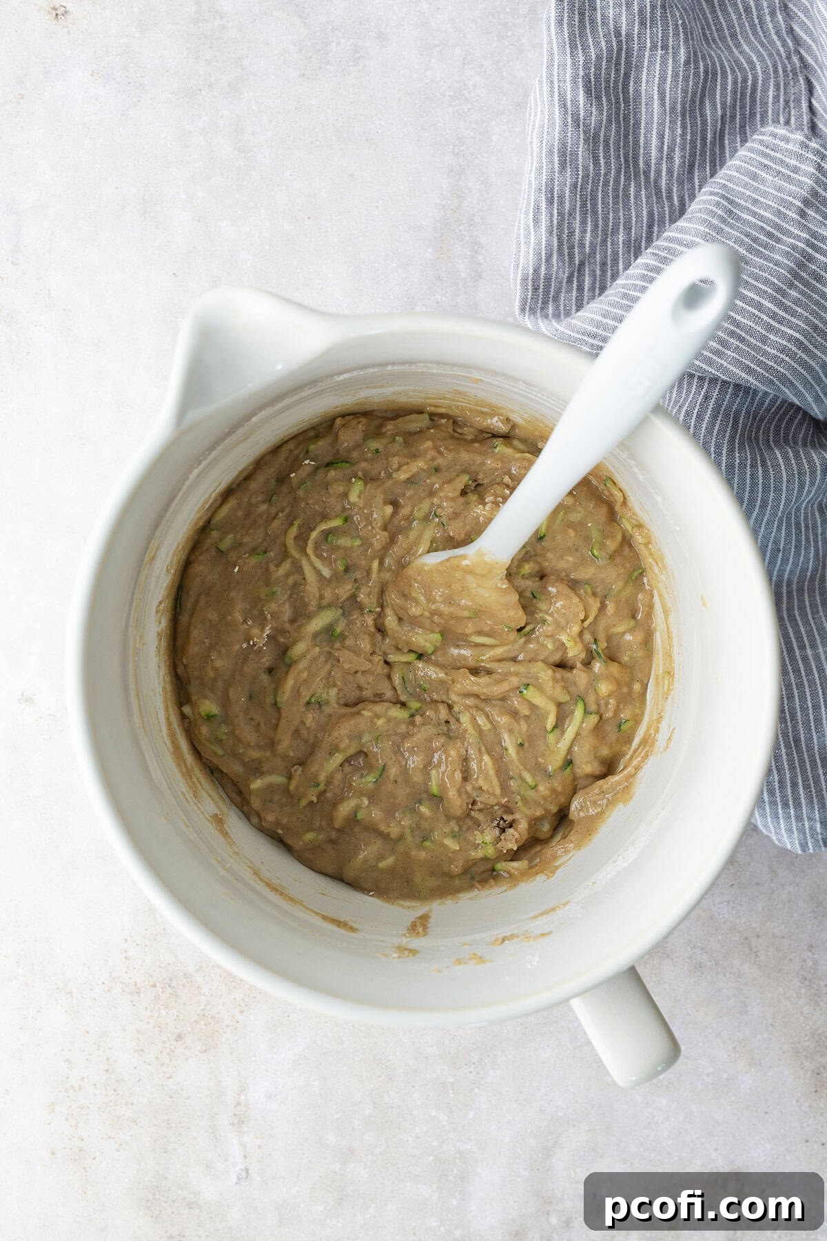 Dry and wet ingredients for zucchini cake combined in a bowl, being gently folded with a rubber scraper.