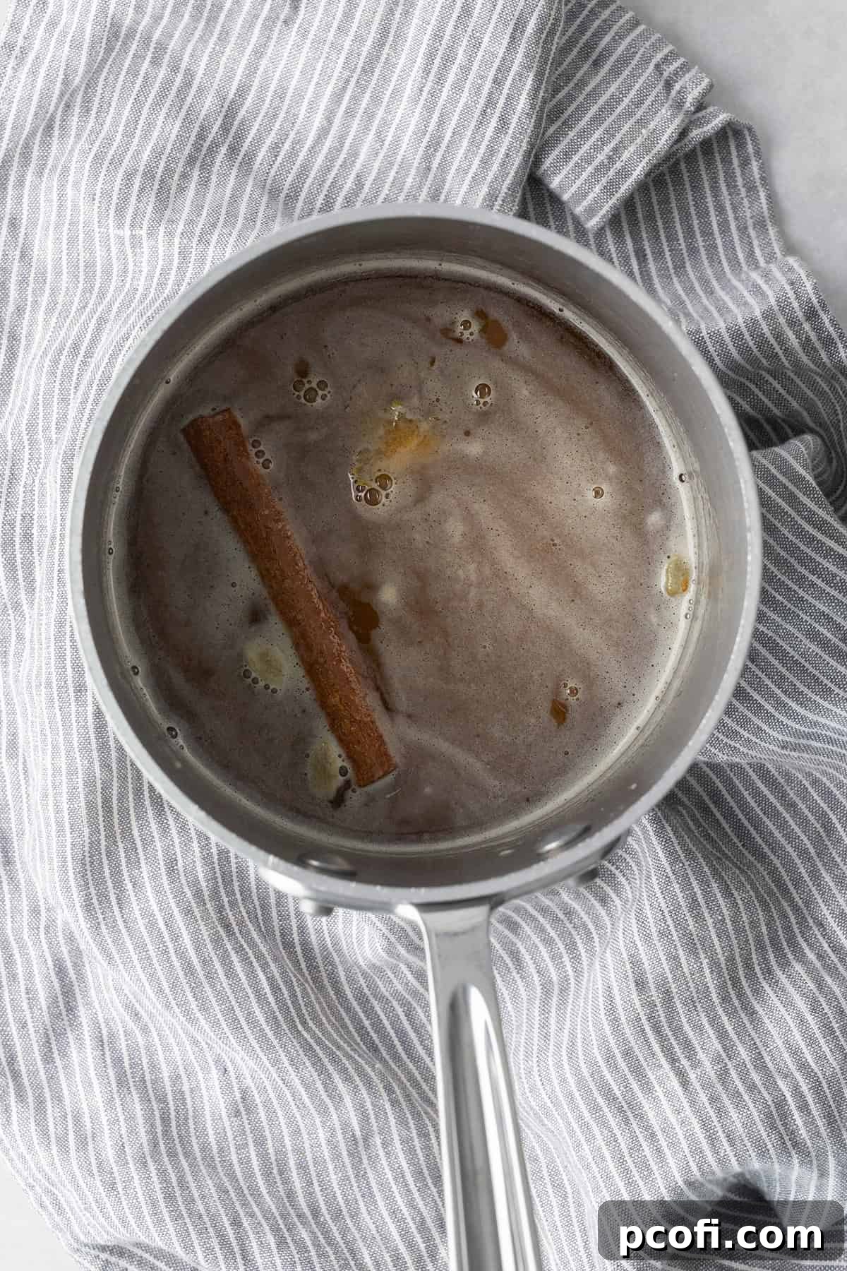 A close-up shot of spiced honey syrup simmering in a silver saucepan, with visible cinnamon sticks and cardamom pods.