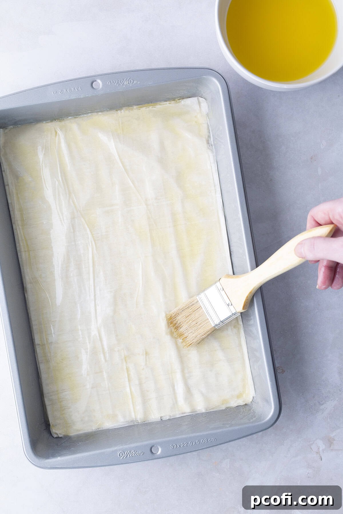 A pastry brush applying melted butter to a folded phyllo dough sheet in a baking pan, showing the meticulous layering process.