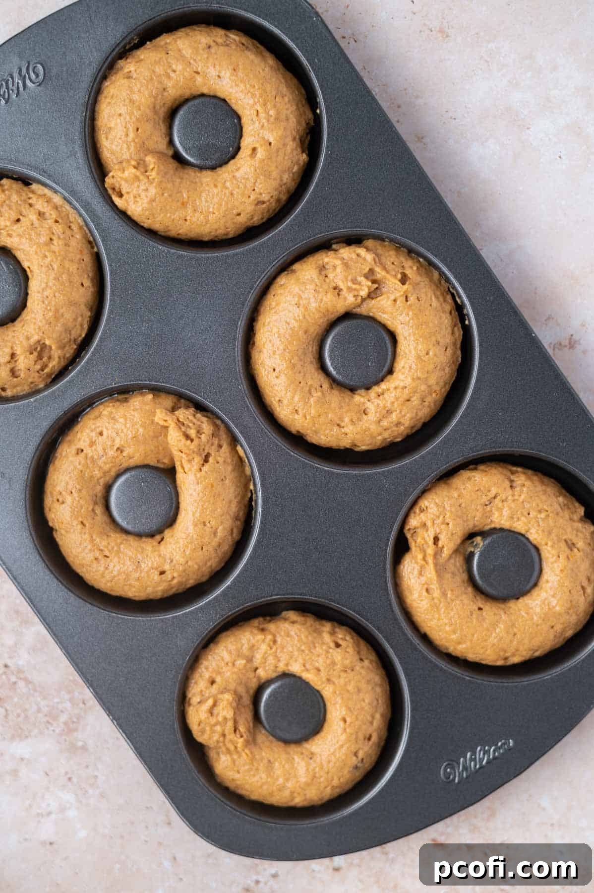 Raw pumpkin donut batter is piped neatly into a greased donut pan, filling each mold evenly.