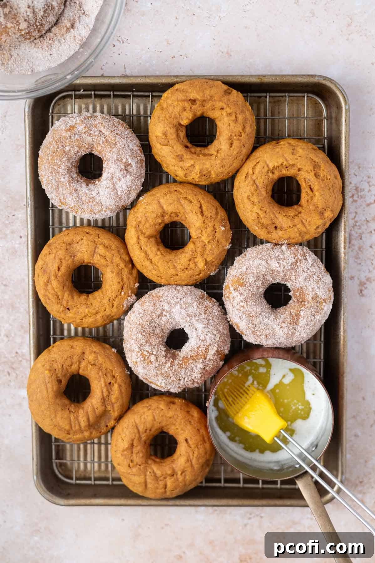 Freshly baked pumpkin donuts on a baking tray, some brushed with melted butter and others dipped in spiced cinnamon sugar.