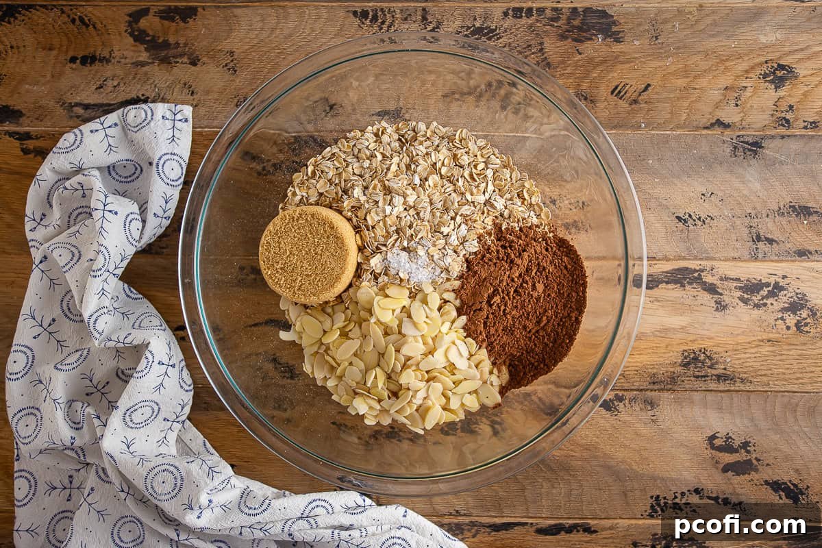 Dry ingredients for chocolate granola in a large glass bowl before being mixed, showing oats, almonds, and cocoa.