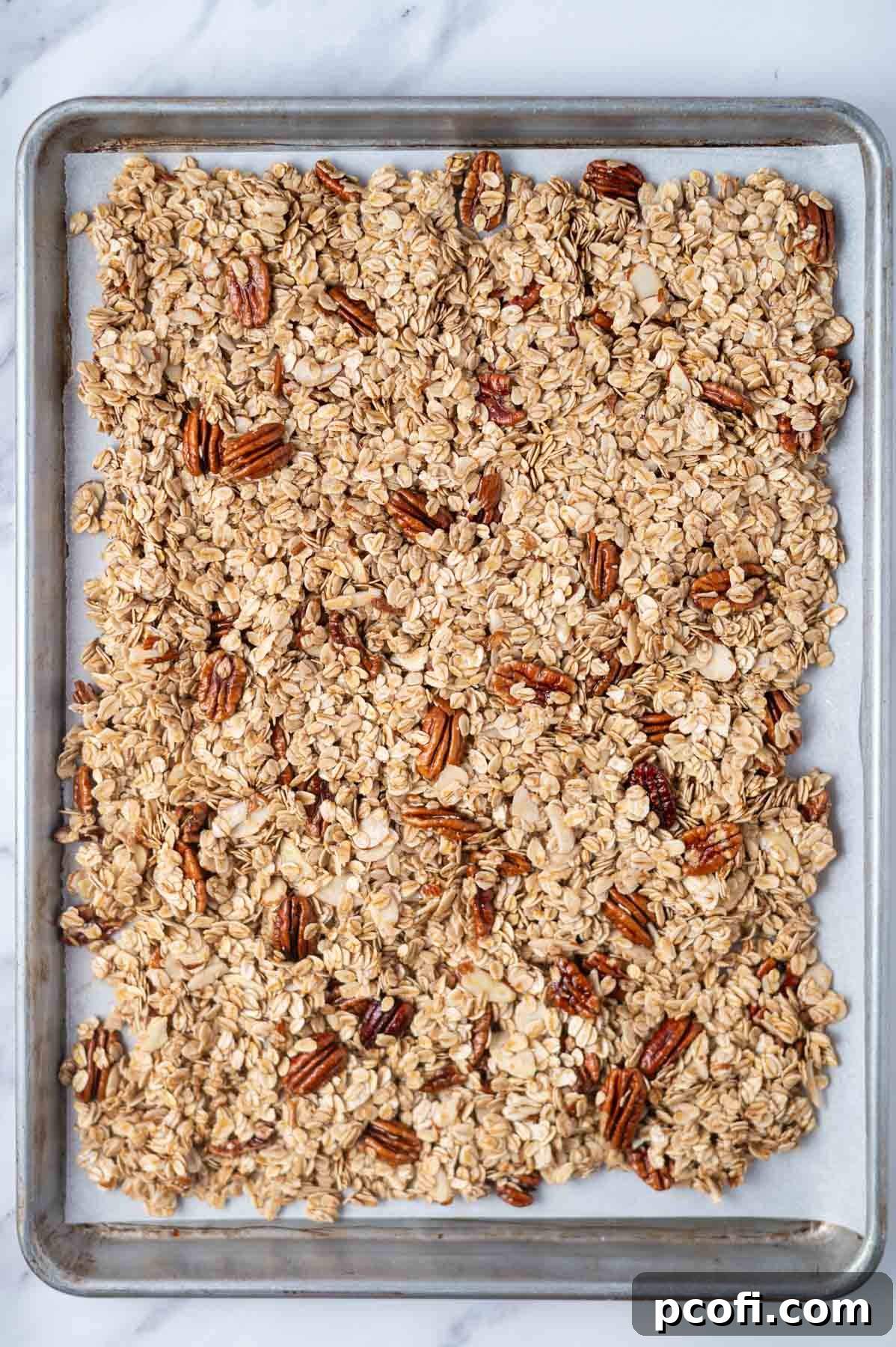 Maple granola spread in a single, even layer on a parchment-lined baking sheet, ready for baking.