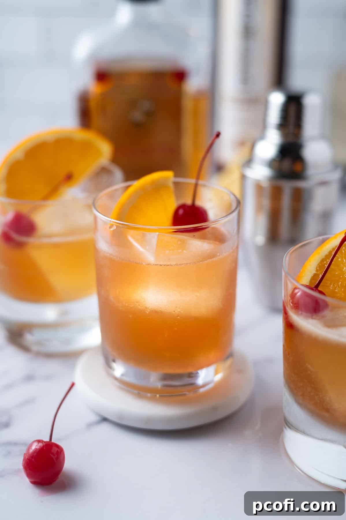 Three beautifully garnished Amaretto Sour cocktails with orange slices and cherries, with bottles of amaretto and bourbon blurred in the background