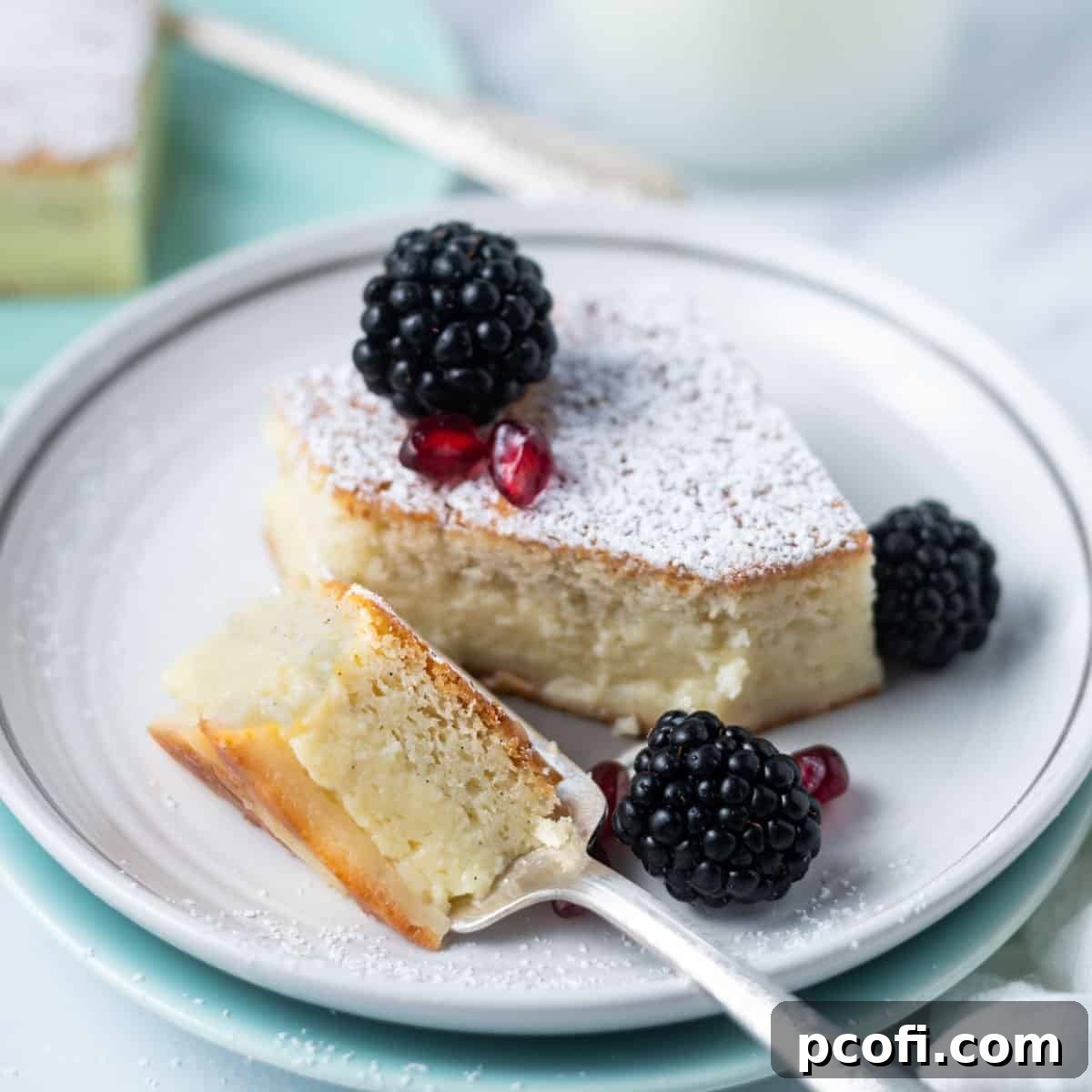 A delectable slice of vanilla bean magic custard cake, garnished with fresh blackberries and vibrant pomegranate arils, a fork poised for a bite