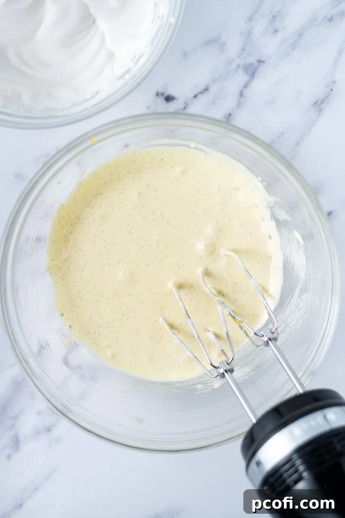 Egg yolk and sugar batter mixed with butter in a glass bowl, being beaten by a hand mixer