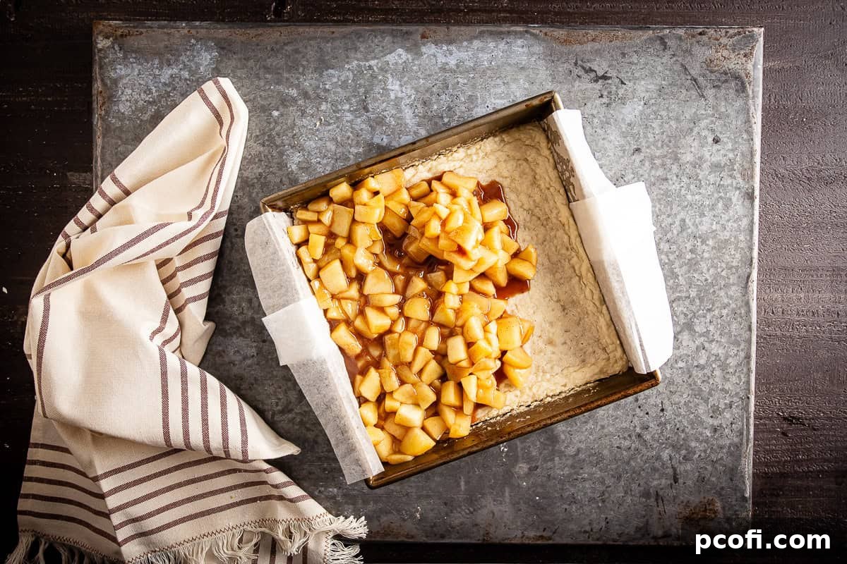 The cooked, fragrant apple pie bar filling being smoothly spread over the pre-baked shortbread crust in the baking pan.