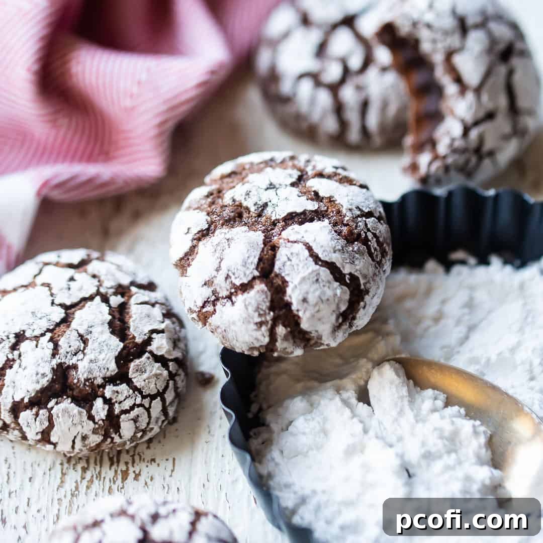 Festive chocolate crinkle cookies, dusted with powdered sugar, presented with a dish of extra sugar and a vibrant red kitchen towel.