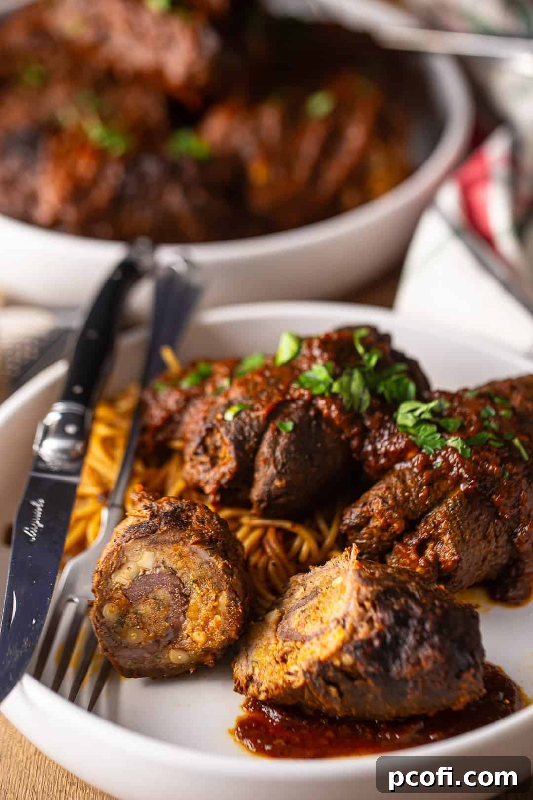 Braciole sliced in half in a large white serving bowl with a fork and knife, garnished with fresh basil.